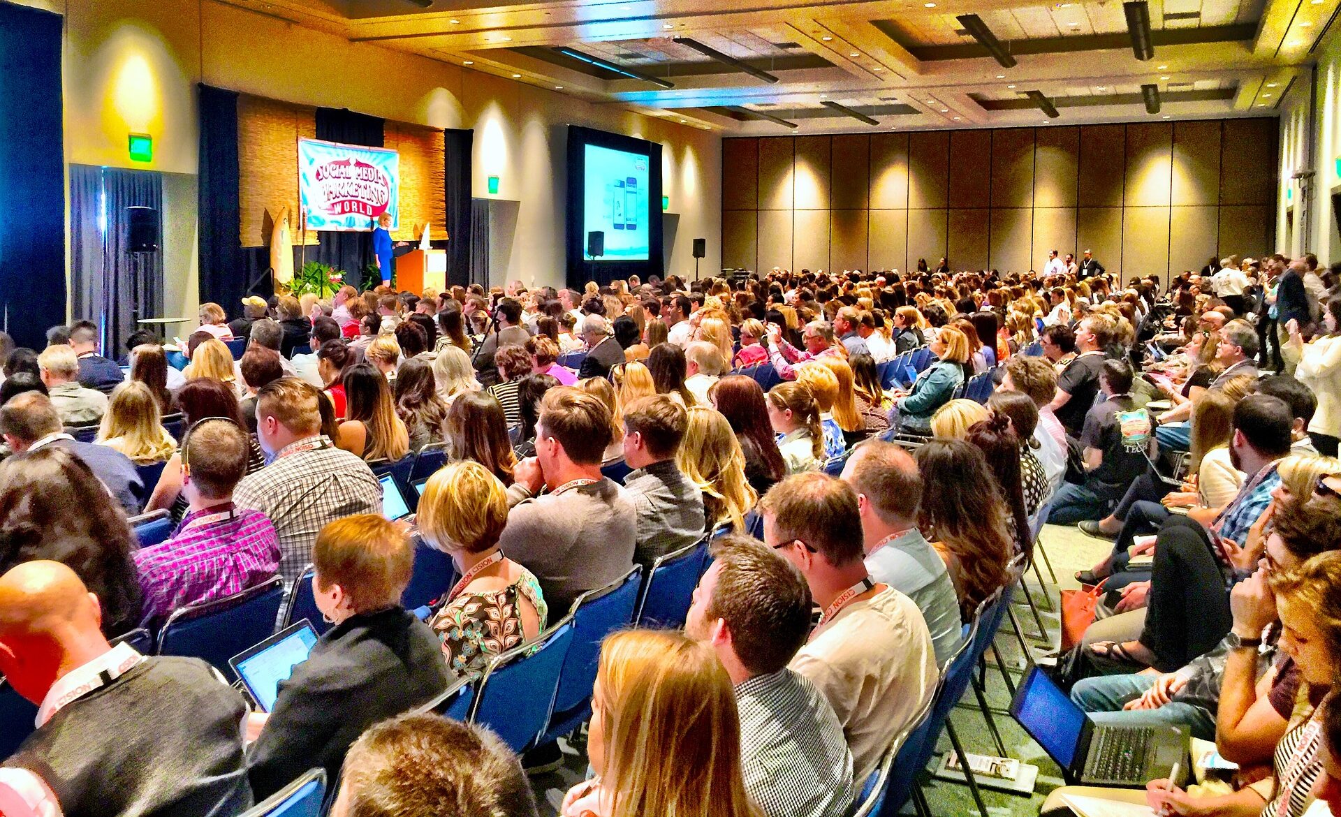 Large audience seated in a conference hall, facing a stage with a presentation screen.