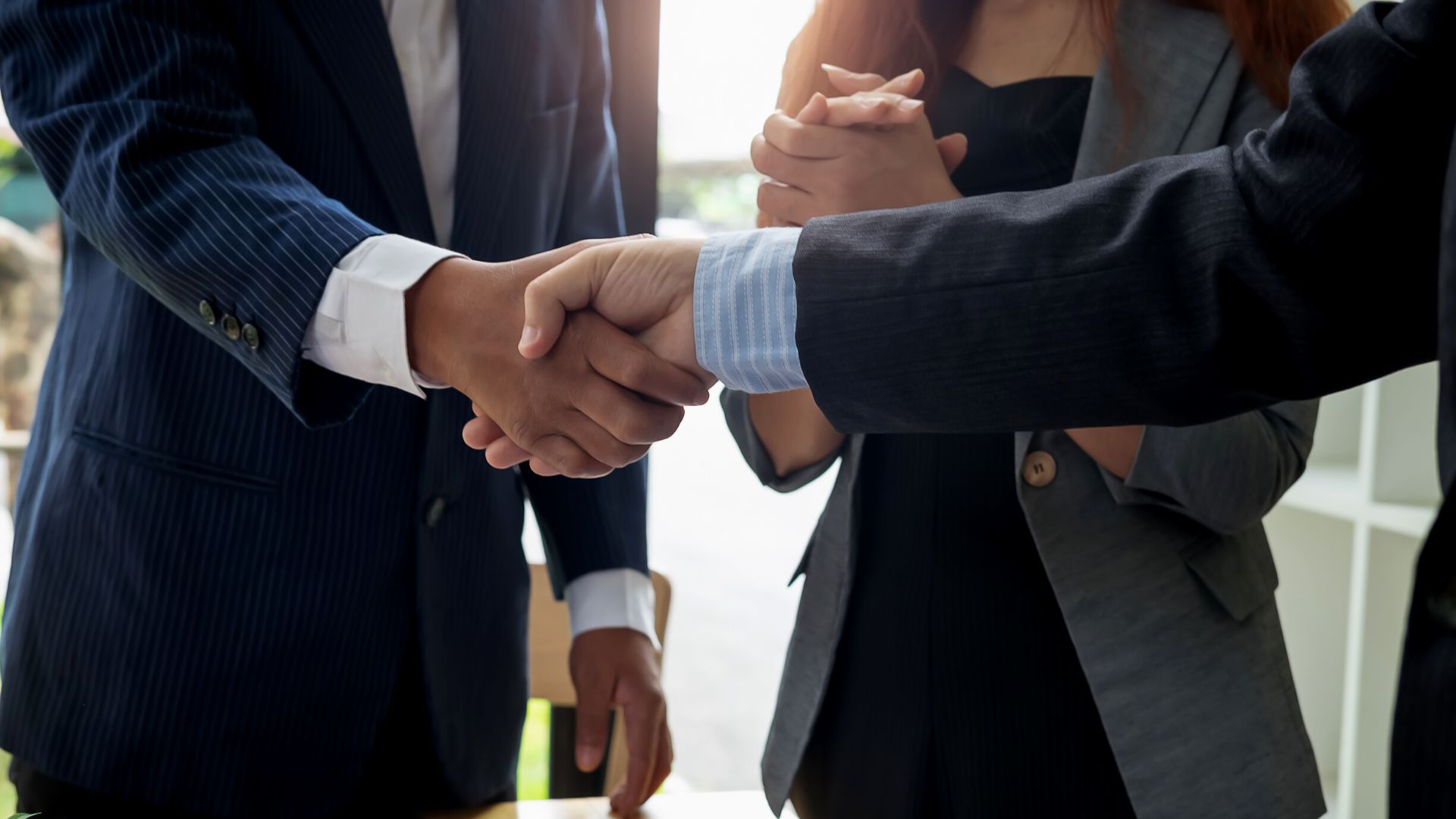 Two business professionals shaking hands in a formal setting, with a third person in the background.